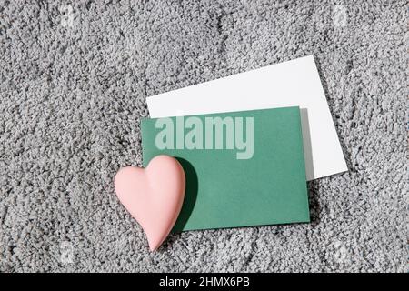 Grußkarte zum Valentinstag. Weiße und grüne Umschläge, Keramik rosa Herz auf einem grauen flauschigen Teppich Hintergrund. Speicherplatz kopieren. Für Text platzieren Stockfoto