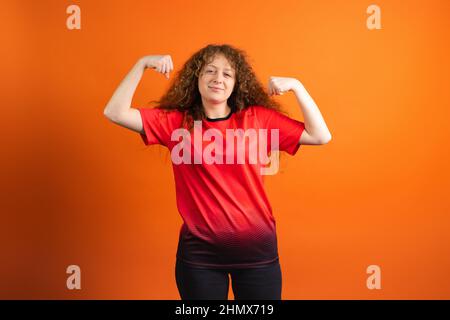 Rothaarige Fan-Fußballfrau im roten T-Shirt, die ihre Muskeln zeigt und ihre Lieblings-Fußballmannschaft bei der Meisterschaft unterstützt. Stockfoto