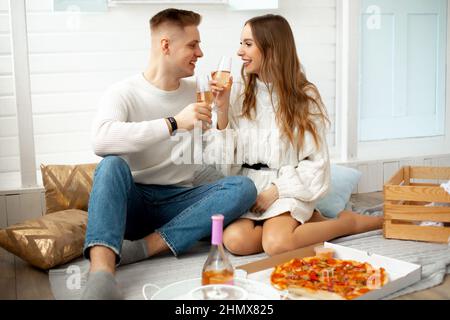 Glücklich zusammen. Schöne junge Leute haben zu Hause ein Date mit Pizza und Wein arrangiert, sie sitzen auf dem Boden gegen die weiße Wand und bringen Glasgläser mit Stockfoto