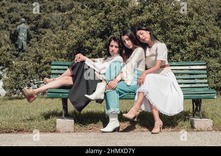 Drei lateinamerikanische Mädchen im Retro-Stil sitzen auf einer grünen Holzbank auf einem platz unter der Sonne mit Bäumen dahinter. Vintage- und Lifestyle-Konzept. Stockfoto