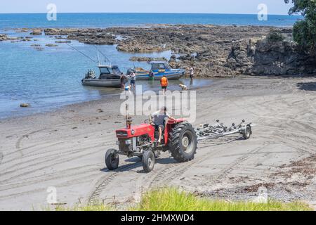 TE Kaha Neuseeland - Januar 31 2022; Boote kehren an Land zurück und werden von einem roten Traktor am Strand zwischen felsigen Gebieten abgeholt. Stockfoto