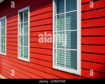 Drei einfache weiße geschlossene Fenster mit Glas- und louvre-Dekoration an der roten Holzdielenwand, Wohn-, Blick von außen. Stockfoto