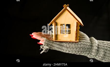 Crop Person mit Holzspielzeug Haus auf schwarzem Hintergrund. Draufsicht auf Ernte anonyme Person hält kleine hölzerne Spielzeughaus auf schwarzem Hintergrund. Stockfoto