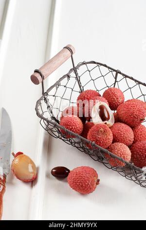 Frische reife Lychee-Früchte und geschälte Lychee auf Drahtkorb, über dem weißen Holztisch. Speicherplatz Kopieren Stockfoto