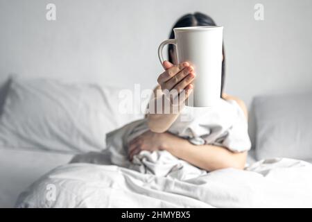 Eine Frau hält eine Tasse Kaffee, während sie im Bett liegt. Stockfoto