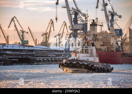 Kleiner eisbedeckter Schlepper im Winter in einem großen Seefrachthafen vor dem Hintergrund von Hafenkranen und Liegeplätzen Stockfoto