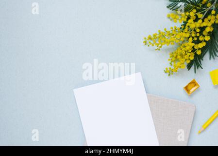 Weißes Blatt Papier für Textkopien, umgeben von Frühlingsmimosen, Notizblock, gelbem Bleistift, Wasserfarben in Küvetten auf blauem Hintergrund. Arbeitsplatz Stockfoto