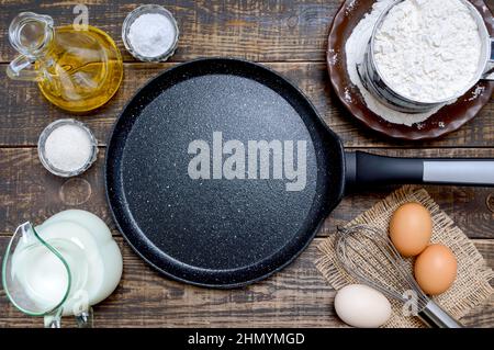 Leere Bratpfanne schwarz mit steinerner Antihaftbeschichtung zum Backen von Pfannkuchen und Zutaten auf Holzhintergrund. Frühstück, Blick von oben. Stockfoto