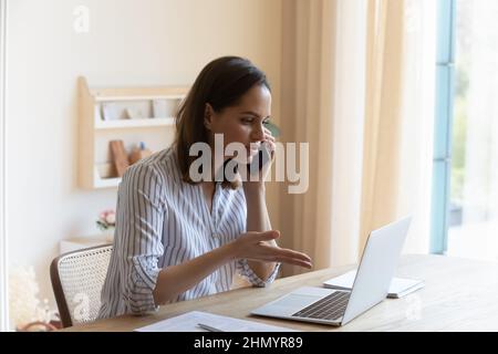 Fokussierte ernsthafte junge Geschäftsfrau, die am Handy spricht und am Computer arbeitet. Stockfoto