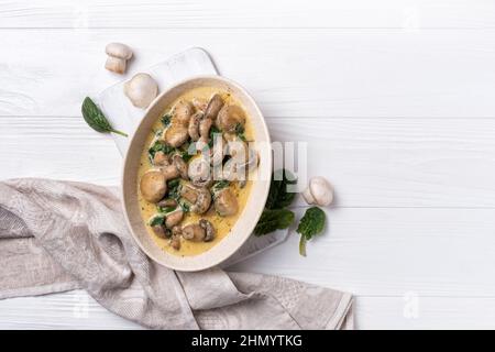 Draufsicht auf Champignon-Pilze in Schüssel mit Spinatblättern und Sahnesauce auf weißem Holzhintergrund Stockfoto