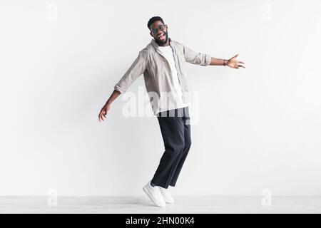 In voller Länge ein cooler junger schwarzer Mann in lässiger Kleidung, der auf Zehenspitzen steht und gegen die weiße Studiowand springen möchte, um Platz zu kopieren Stockfoto