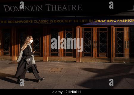 Eine Frau, die eine Gesichtsmaske, eine lange Jacke, einen Schal und eine Hose trägt, kommt weit vorbei am Londoner Dominion Theatre Stockfoto