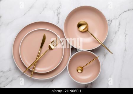 Geschirr und Utensilien zum Servieren und Essen von Mahlzeiten. Beigefarbene runde Teller und goldfarbenes Besteck auf einem weißen Marmortisch, Draufsicht. Moderne Keramik Stockfoto