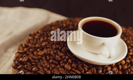 Tasse Kaffee und Untertasse auf Kaffeebohnen. Zusammensetzung aus frisch gebrühtem Kaffee auf gerösteten Bohnen. Konzept der Zubereitung duftendes Getränk Stockfoto