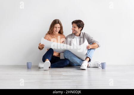 Fröhliche europäische Millennial Dame und Frau sitzen auf dem Boden in leeren Raum Blick auf den Hausplan und Planung neuer Innenausstattung Stockfoto