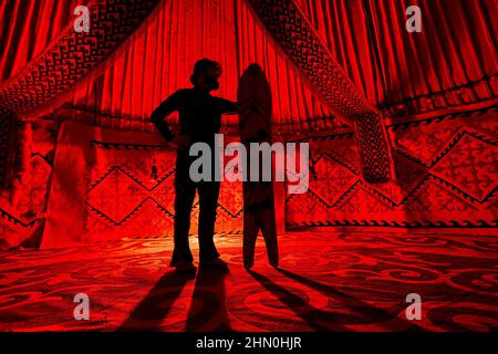 Silhouette von man Holding mit gespaltenem Schneeboard im Yurt Nomadenhaus gegen Teppich mit ethnischen Mustern, die von einem roten Licht im Inneren beleuchtet werden. Backcountry r Stockfoto