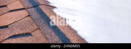 Rote Gürtelrose. Das Dach eines Hauses aus Schindeln ist im Winter mit Schnee bedeckt, Nahaufnahme. Frostbeständigkeit und Elastizität von bituminösen Fliesen in Stockfoto