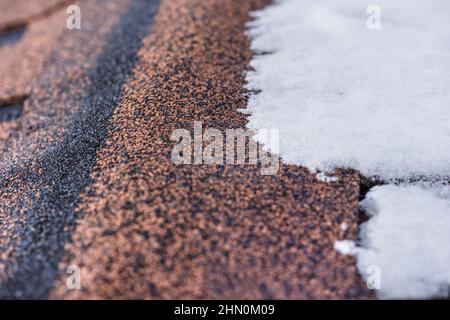 Rote Gürtelrose. Das Dach eines Hauses aus Schindeln ist im Winter mit Schnee bedeckt, Nahaufnahme. Frostbeständigkeit und Elastizität von bituminösen Fliesen in Stockfoto