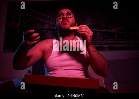 Ein 40-jähriger, unrasiert übergewichtiger Mann schaut Fernsehen, zeigt verschiedene Emotionen und isst Pizza. Fauler Mann mittleren Alters. Stockfoto