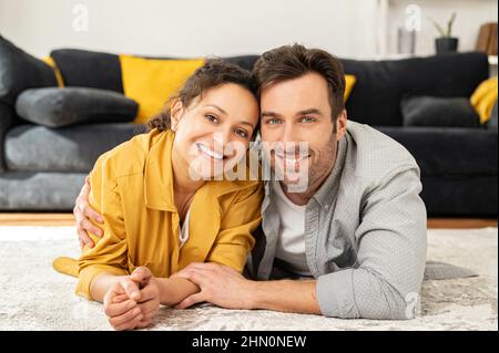 Ruhiges und unbeschwertes Paar verliebt in die Kamera und lächelnd. Gutaussehender Mann mittleren Alters und charmante Frau, die sich umarmt auf dem Boden im Wohnzimmer niederlegt. Liebe und Bindung Konzept Stockfoto