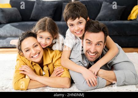 Porträt einer glücklichen multirassischen Familie zu Hause. Multiethnische Ehepartner, ihre niedliche Kleinkindtochter und ihr Sohn im Schulalter blicken in die Kamera und lächeln toothy auf dem Boden liegend Stockfoto