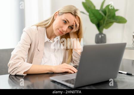 Gelangweilte weibliche Angestellte, die im Büro vor einem Laptop am Schreibtisch sitzt, wegschaut und sich traurig fühlt. Nachdenkliche junge Frau hat keine Inspiration für die Arbeit, Gefühl von Schlafmangel und Müdigkeit Stockfoto