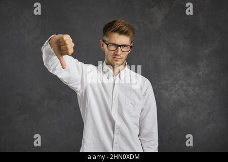 Unzufriedener und frustrierter junger Mann, der mit dem Daumen auf grauem Hintergrund steht. Stockfoto