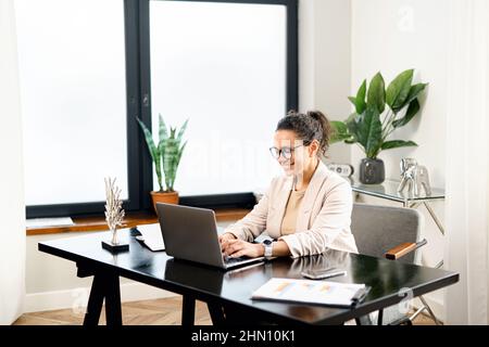 Fröhliche weibliche Angestellte arbeitet mit einem Laptop im modernen Büro. Eine positive Frau mittleren Alters in eleganter Freizeitkleidung, die auf der Tastatur tippt, schreibt eine Journalistin einen Artikel Stockfoto