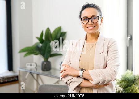 Fröhliche Frau Unternehmer steht mit gekreuzten Armen im zeitgenössischen Büro, multirassische freiberufliche Frau in eleganter Freizeitkleidung blickt auf die Kamera mit gekreuzten Armen, Kleinunternehmer Stockfoto