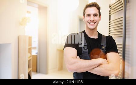 Lächelnder, hübscher Klempner, der mit gekreuzten Armen steht und die Kamera im Badezimmer anschaut Stockfoto