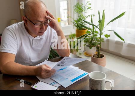 Ein älterer Mann, der seinen Kopf hält und versucht, die anhebenden Kosten für Energie- und Steuerrechnungen zu bewältigen. Stockfoto