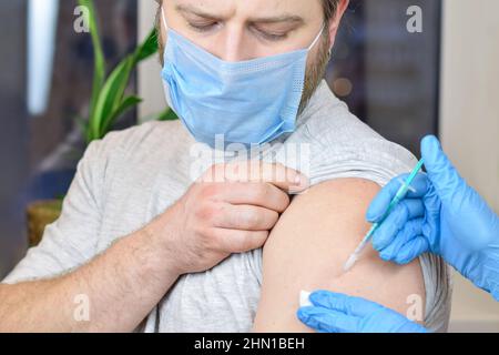 Hände eines Arztes, der Coronavirus-Covid-19-Impfstoff in eine Impfspritze in den Armmuskel des kaukasischen Mannes zur Covid-Immunisierung injiziert. Stockfoto