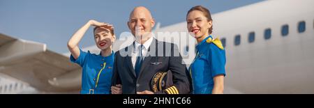 Glücklicher Pilot und zwei attraktive Stewardessen stehen vor einem Flugzeug und lächeln nach der Landung Stockfoto