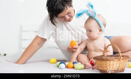 Mutter und Kind mit bunten Eiern. Mama und Baby mit Hasenohren. Eltern und Kinder spielen im Frühling drinnen. Familie feiert Ostern Stockfoto