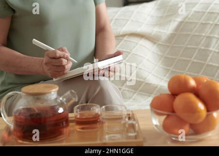 Beschnittenes Bild einer Frau, die Tee trinkt und eine Einkaufsliste aufschreibt, wenn sie auf dem Sofa im Zimmer sitzt Stockfoto