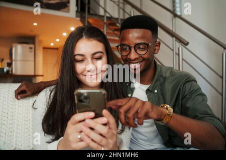 Afroamerikanischer Mann, der mit einer kaukasischen Freundin auf einem Mobiltelefon sitzt, während er auf dem Sofa sitzt Stockfoto