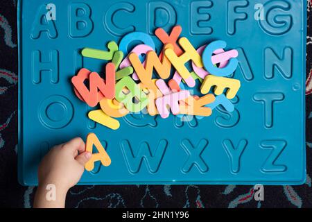 Ein kleines Kind Hand Sortieren der pädagogischen bunten schwammigen Buchstaben in gravierten blauen Blatt, oben Standpunkt Stockfoto