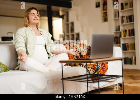 Junge Schwangere meditieren und verwenden Laptop, während sie zu Hause auf der Couch sitzen Stockfoto