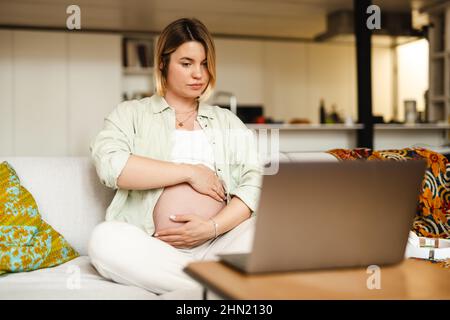 Eine Schwangere zeigt ihren Bauch und benutzt einen Laptop, während sie zu Hause auf der Couch sitzt Stockfoto