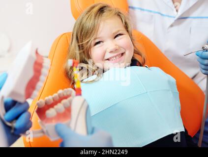 Die Kinderzahnärztin zeigt einem kleinen Mädchen, das in einem orangen Zahnstuhl sitzt und lächelt, wie man richtig ihre Zähne putzt. Stockfoto