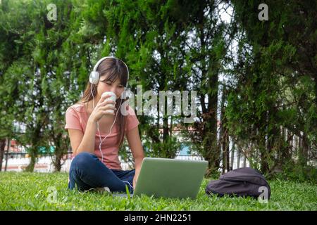 High School asiatische Teenager Mädchen entspannen auf dem Gras in einem öffentlichen Park mit Laptop liegen und Musik hören Stockfoto