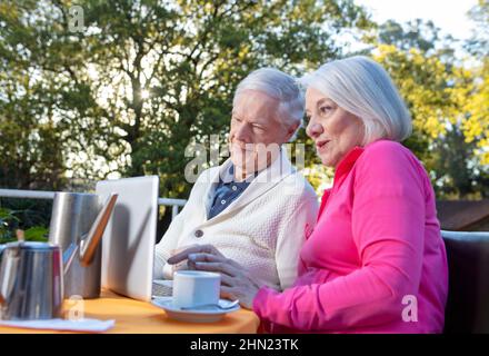Ältere Menschen leben ihr Leben von seiner besten Seite. Ein paar Rentner, die draußen frühstücken und einen Laptop benutzen Stockfoto