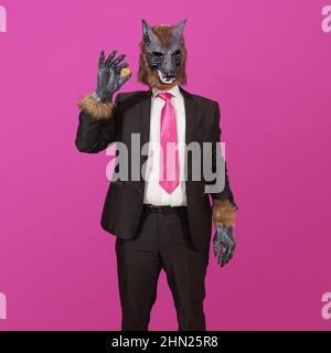 Auf einem rosa Hintergrund ist ein Mann in einem schwarzen Anzug mit einer Jacke, einem weißen Hemd und einer Krawatte, der eine Werwolf-Maske trägt und eine physische Darstellung trägt Stockfoto