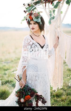 Die Braut in einem Kranz steht in der Nähe des Hochzeitsbogens auf der Wiese Stockfoto