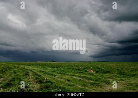 Ein vorhersehender, dramatischer, dunkelgrauer Gewitter wolkelt den Himmel über einem offenen Grasflugplatz und grünen Wiesen Stockfoto