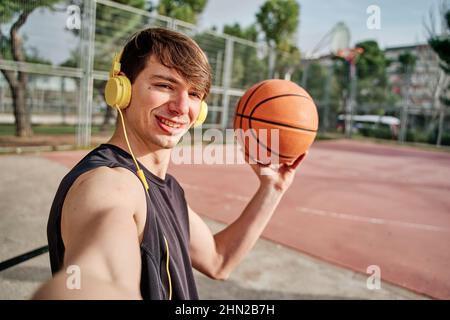 Junger Athlet, der ein Selfie mit gelben Kopfhörern gemacht hat. Hält einen Basketball Stockfoto