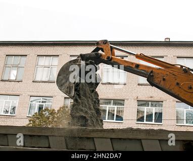 Straßenarbeiten auf der Stadtstraße. Der Baggereimer sammelt den alten Gehweg und lädt ihn in einen Muldenkipper. Stockfoto