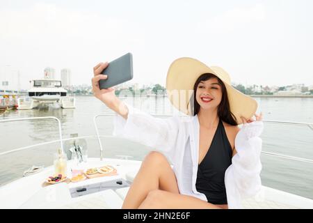 Lächelnde junge Frau im weißen Hemd über Badeanzug und Strohhut beim Selfie beim Entspannen auf luxuriösen kleinen Jachten Stockfoto