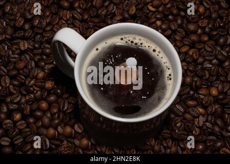 Das Bild zeigt eine Tasse Kaffee mit einem herabfallenden Milchtropfen. Im Hintergrund sind Kaffeebohnen zu sehen. Stockfoto