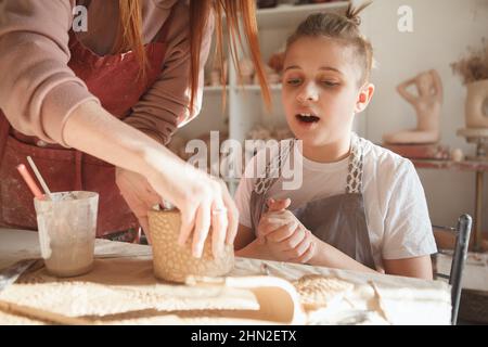 Der junge Junge schaut aufgeregt und sieht einem professionellen Keramiker zu, der ihm beim Töpferunterricht beim Tee-Krug hilft Stockfoto